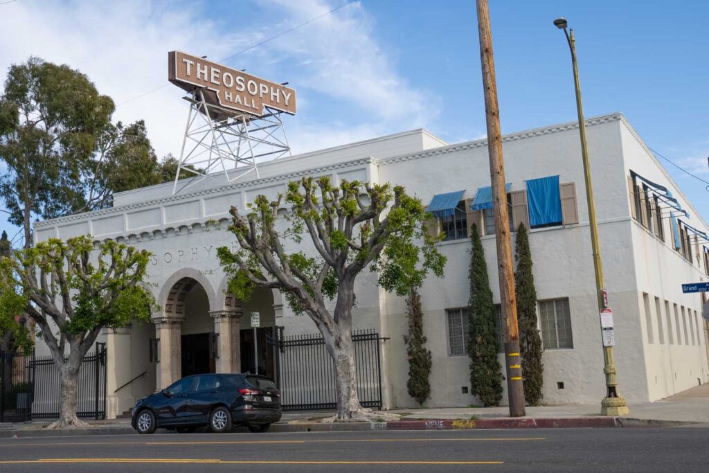 Wide-angle street-view of Theosophy Hall’s white stucco façade, trimmed trees, and rooftop sign reading “THEOSOPHY HALL.”