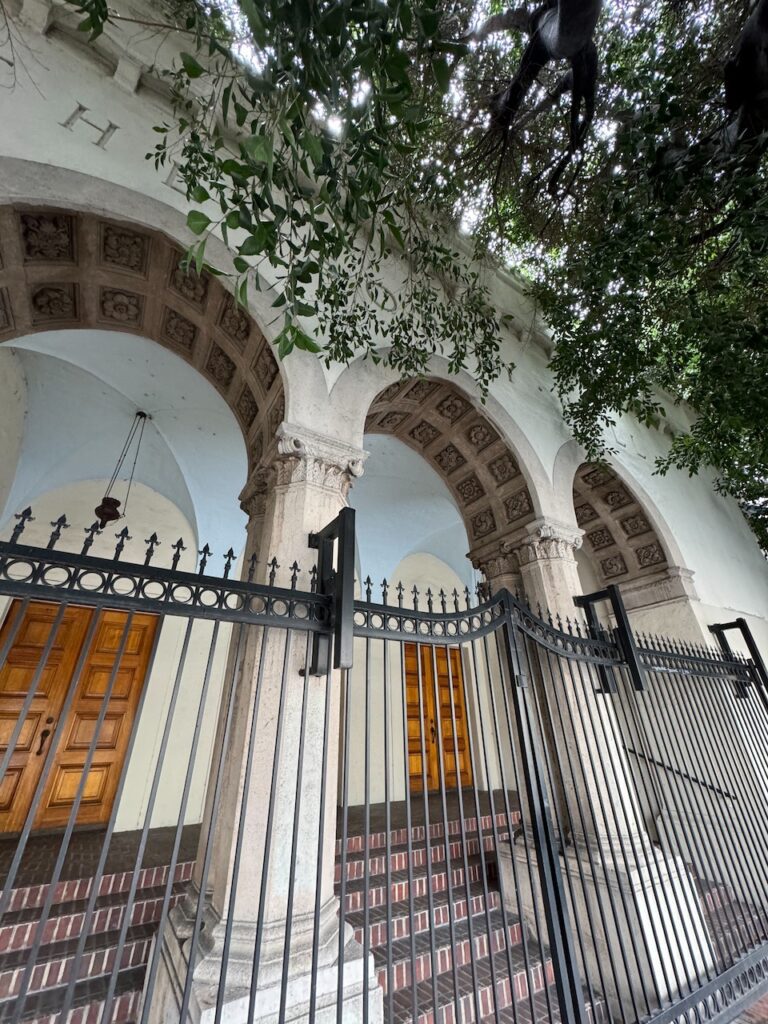 Wrought-iron fence in front of a stucco portico with carved stone arches and wooden entry doors, framed by overhanging tree branches.