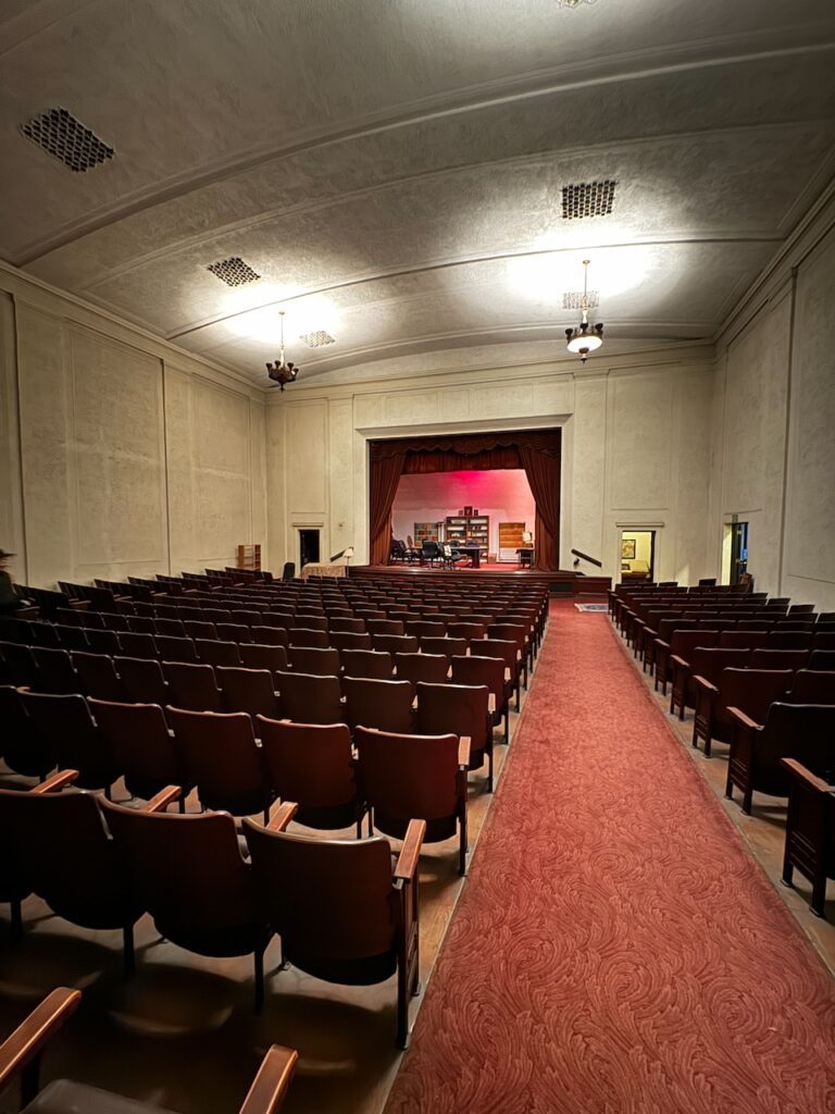 Interior view of Theosophy Hall auditorium with rows of wooden seats flanking a central red-carpeted aisle leading to a stage framed by a proscenium arch.