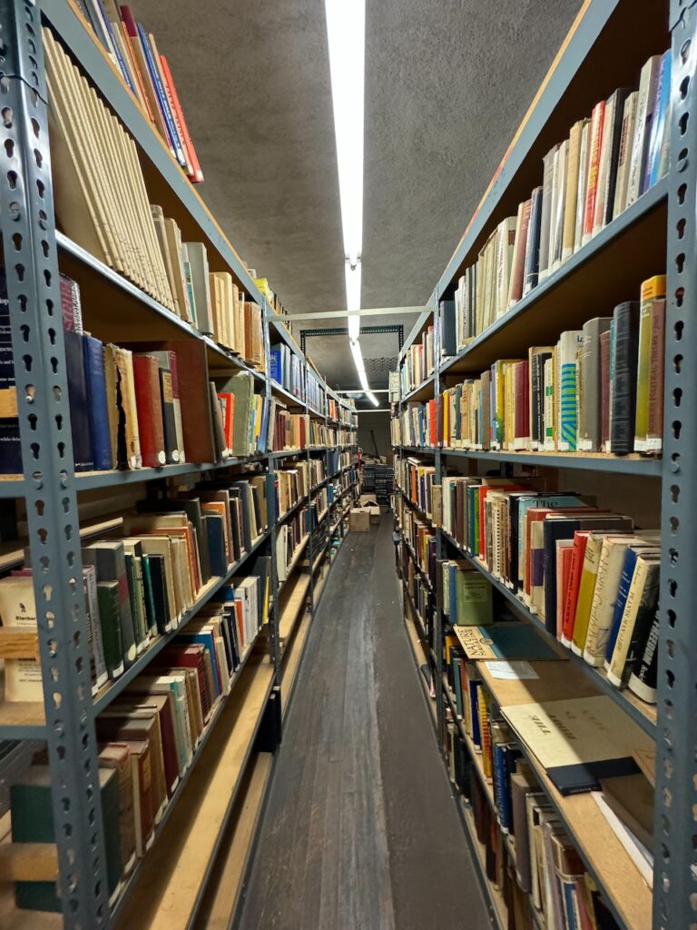 Narrow aisle between tall metal bookshelves packed with assorted hardcover volumes under fluorescent lighting.