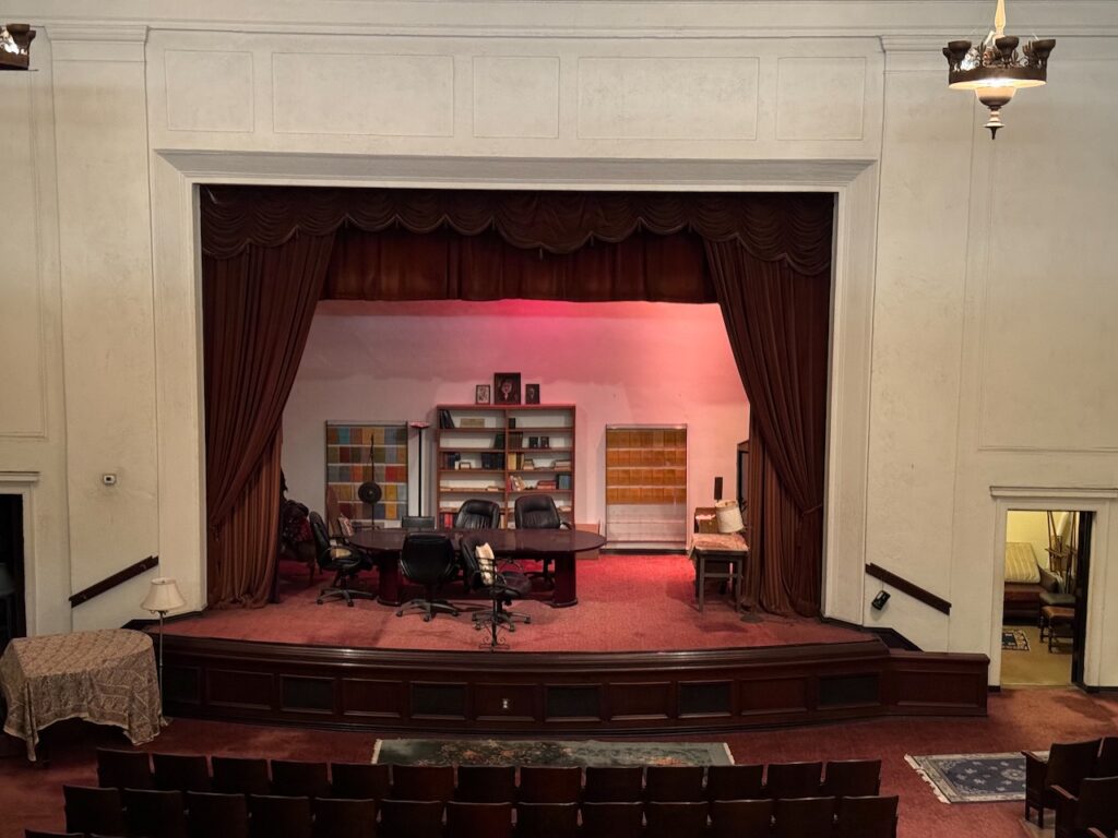 Proscenium stage at Theosophy Hall set with a conference table, rolling chairs, bookshelves, and red velvet curtains.